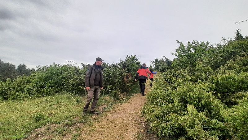 Na zdjęciu widzimy grupę osób poruszających się ścieżką wśród gęstej zieleni. Na pierwszym planie znajduje się mężczyzna w mundurze, najprawdopodobniej pracownik służby ochrony przyrody lub leśnictwa — można zauważyć naszywkę na rękawie jego kurtki. W tle, na ścieżce, widzimy trzy osoby, ubrane w czerwone kurtki, idące w stronę lasu lub zalesionego wzgórza. Scena ma miejsce w terenie o pagórkowatym krajobrazie.