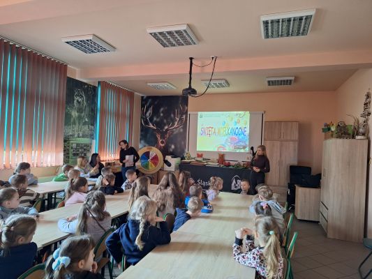 Zdjęcie pokazuje dzieci siedzące w Sali edukacyjnej. Pracownik ZPKWŚ opowiada o świętach wielkanocnych i wyświetla prezentację multimedialną z napisem „Święta Wielkanocne”.