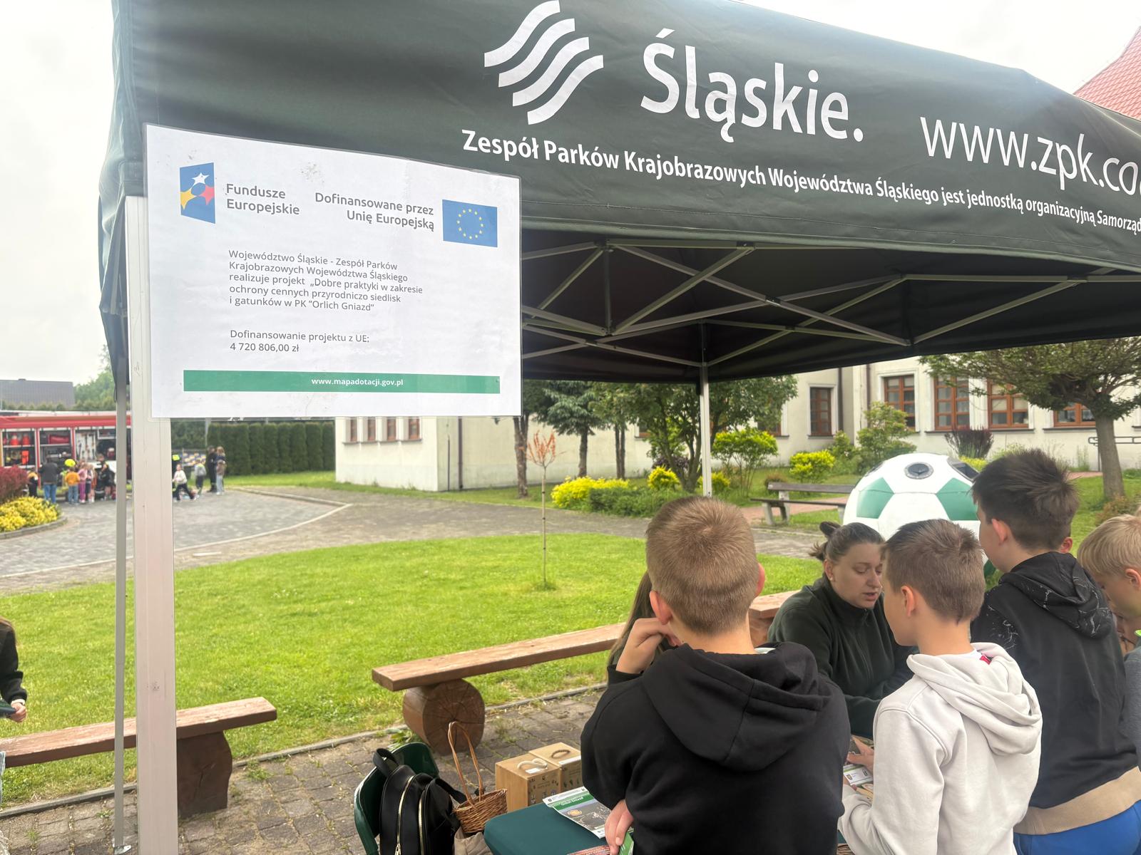 Na zdjęciu widać plenerowe stoisko edukacyjne pod zielonym namiotem z napisem „Śląskie. Zespół Parków Krajobrazowych Województwa Śląskiego”. Pod namiotem grupa dzieci rozmawia z prowadzącą zajęcia kobietą. Na pierwszym planie po lewej stronie umieszczono białą tablicę informacyjną o projekcie unijnym realizowanym w Parku Krajobrazowym „Orlich Gniazd” (kwota dofinansowania: 4 720 806,00 zł). W tle widoczny jest trawnik, budynki oraz drewniane ławki.