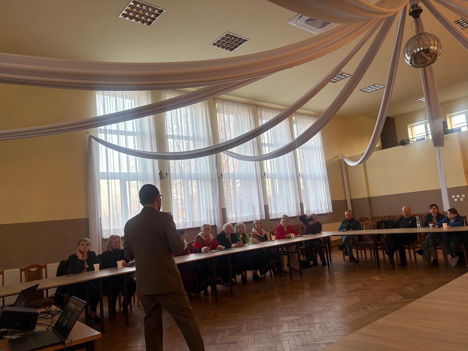 Mężczyzna stojący na środku sali przemawia do grupy osób siedzących przy stołach ustawionych w kształcie podkowy. Spotkanie odbywa się w jasnej sali z dużymi oknami,  a z sufitu zwisają dekoracyjne tkaniny i kula dyskotekowa. Na stole obok stoi laptop używany do prezentacji.