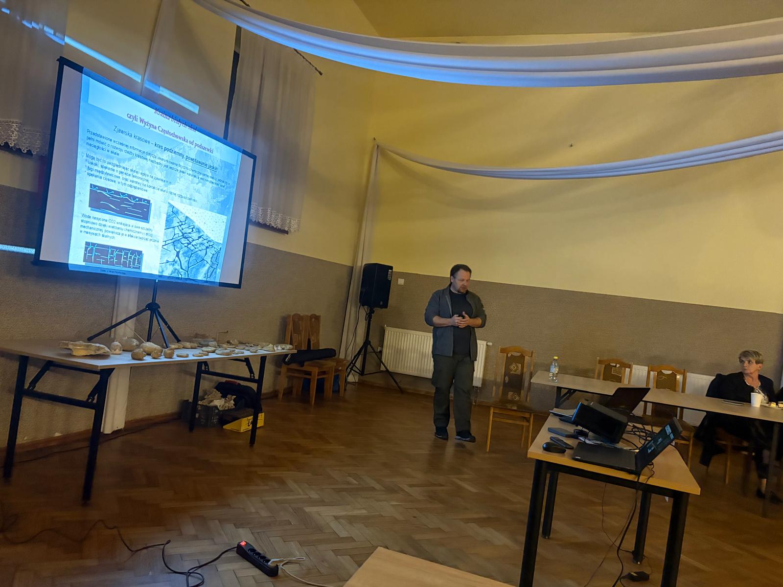 W sali konferencyjnej mężczyzna prowadzi prezentację z projektora dotyczącą zjawisk krasowych. Na ekranie widoczny jest slajd z diagramami i tekstem, a obok na stole leżą różne okazy skał. Przy stołach siedzą ludzie i słuchają wykładu.