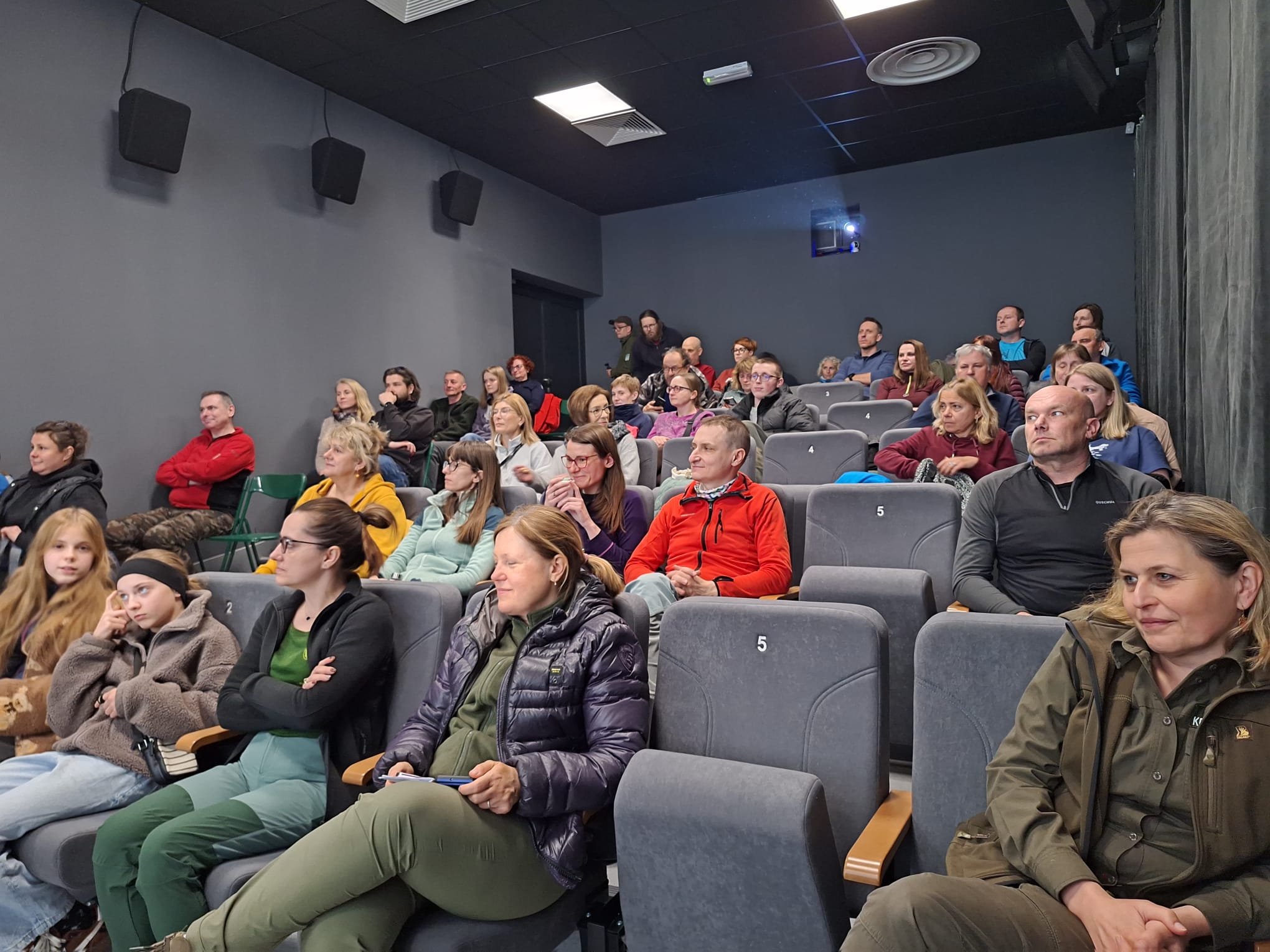 Grupa kilkudziesięciu osób w różnym wieku siedzi w rzędach szarych foteli w niewielkiej sali kinowej. Wszyscy są zwróceni w jedną stronę i uważnie obserwują coś poza kadrem, prawdopodobnie ekran lub prezentację. W pomieszczeniu panuje stonowane oświetlenie, a na suficie widać projektor oraz system wentylacji. Uczestnicy mają na sobie casualowe, często outdoorowe ubrania, takie jak kurtki i bluzy