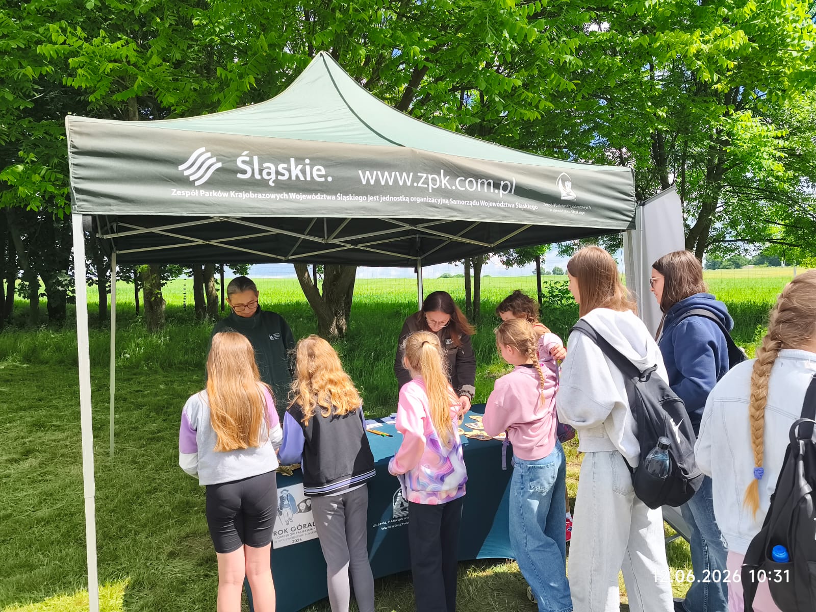 Plenerowe stoisko edukacyjne Zespołu Parków Krajobrazowych Województwa Śląskiego (ZPKWŚ) podczas pikniku w Smoleniu. Pod ciemnozielonym namiotem z białym logo „Śląskie.” oraz adresem strony „www.zpk.com.pl” dwie edukatorki rozmawiają z grupą dzieci i młodzieży stojącą wokół stołu. Na stole znajdują się materiały warsztatowe, a na przedniej płachcie obrusu widnieje grafika z napisem „Rok Górali 2026”. W tle rozciąga się zielona polana i drzewa.