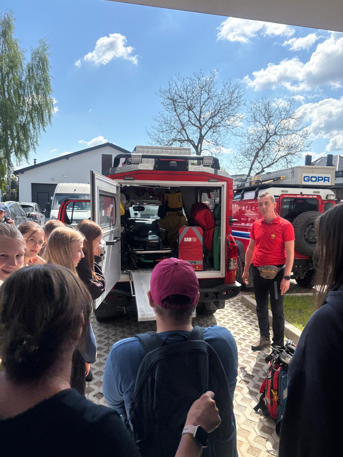 Zdjęcie przedstawia prezentację specjalistycznego sprzętu ratowniczego dla grupy dzieci  i młodzieży na świeżym powietrzu w słoneczny dzień. Głównym elementem jest otwarty tył czerwonego samochodu terenowego GOPR, w którym widoczne jest wyposażenie medyczne  i ratunkowe, w tym nosze, torby i liny. Obok pojazdu stoi ratownik górski w czerwonej koszulce polo i czarnych spodniach, który opowiada o pracy i sprzęcie. Zgromadzona wokół młodzież  z zaciekawieniem zagląda do wnętrza samochodu. W tle widać drugi pojazd ratowniczy  z napisem „GOPR”, budynki oraz drzewa pod błękitnym niebem.