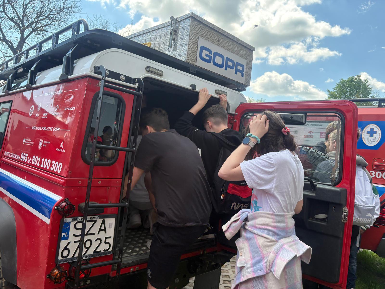 Zdjęcie przedstawia grupę młodzieży oglądającą z bliska terenowy samochód ratowniczy GOPR podczas pokazów na świeżym powietrzu. Pojazd jest czerwony z białym dachem, na którym zamontowano skrzynię z widocznym napisem „GOPR”, a na jego boku i tablicy rejestracyjnej znajdują się oznaczenia i numery alarmowe. Kilku chłopców zagląda do otwartego wnętrza bagażnika samochodu, a na pierwszym planie po prawej stronie stoi dziewczyna w jasnej koszulce, tyłem do obiektywu, obserwująca sytuację. W tle widać fragment drugiego pojazdu ratowniczego oraz błękitne niebo z białymi chmurami.