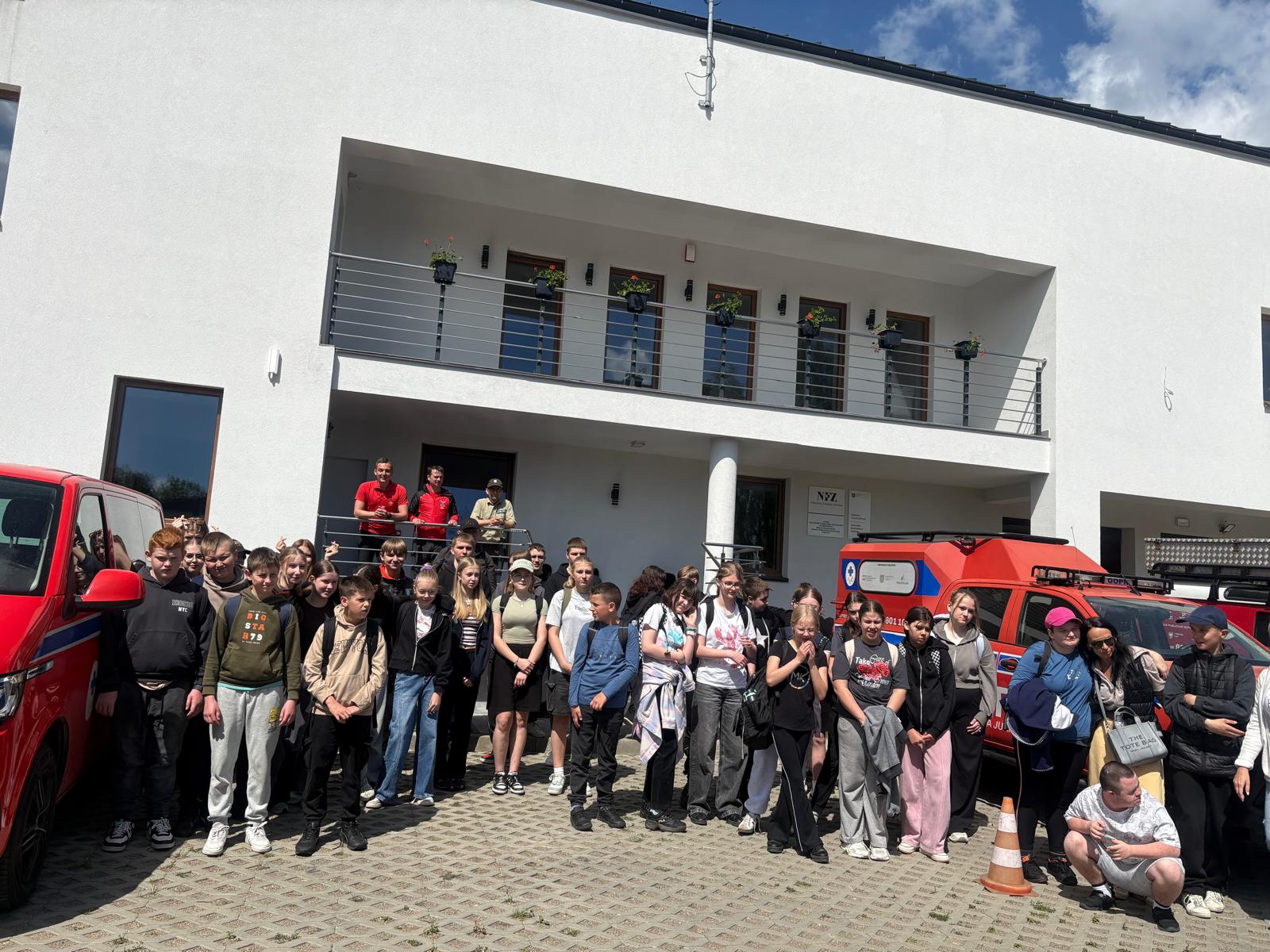 Zdjęcie przedstawia dużą grupę dzieci, młodzieży oraz dorosłych pozujących do wspólnej fotografii pamiątkowej na wybrukowanym placu przed nowoczesnym, białym budynkiem  z balkonem. Uczestnicy spotkania stoją ustawieni w kilku rzędach, a po obu stronach grupy widoczne są czerwone, specjalistyczne samochody ratownicze. W tle, na schodach prowadzących do budynku, stoi trzech mężczyzn, w tym ratownicy w charakterystycznych czerwonych strojach. Całość sceny rozgrywa się w słoneczny dzień pod błękitnym niebem  z białymi chmurami.