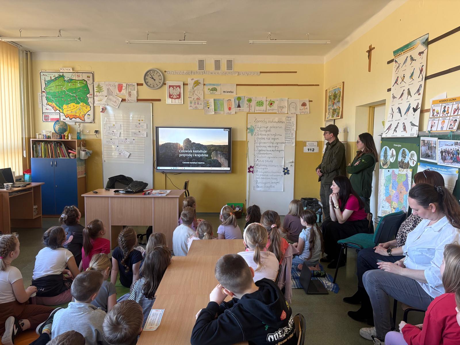 Dzieci siedzą w szkolnej klasie na podłodze i przy ławkach, oglądając film edukacyjny wyświetlany na ekranie. Na ekranie widać slajd dotyczący wpływu człowieka na przyrodę i krajobraz. Z tyłu sali stoją prowadzący zajęcia oraz nauczyciele, a na ścianach znajdują się mapy, plakaty edukacyjne i prace uczniów.